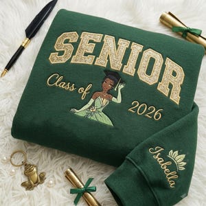 Op de afbeelding: Een donkergroene sweater met de gouden glittertekst SENIOR, de geborduurde tekst Class of 2026 en een afbeelding van een personage in een groene jurk met een afstudeerhoed. De mouw is geborduurd met de naam Isabella en een lotus-icoon.