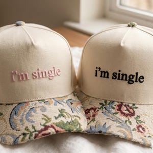 Op de afbeelding: Twee beige baseballpetten met de geborduurde tekst "I'm single". De petten hebben een bloemmotief in blauw, groen en rood. Eén pet heeft roze tekst en de andere heeft zwarte tekst.