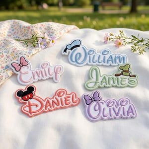 Può includere: Cinque toppe ricamate con nomi e motivi ispirati a Disney. Le toppe mostrano i nomi Emily in rosa con un fiocco, William in blu con un cappello da marinaio, James in verde con un cappello, Daniel in rosso con orecchie e Olivia in viola con un fiocco.