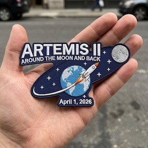 Op de afbeelding: Een marineblauwe patch met de tekst "ARTEMIS II" en "AROUND THE MOON AND BACK". De patch toont een raket, de aarde, de maan en sterren. De datum "April 1, 2026" is ook opgenomen.