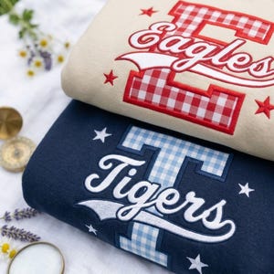 May include: A beige sweatshirt featuring a red and white gingham letter E with the word Eagles in white script, and a navy blue sweatshirt featuring a light blue and white gingham letter T with the word Tigers in white script, both with stars.