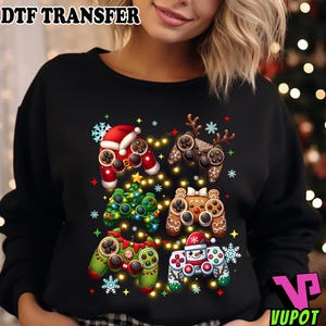 May include: Black sweatshirt featuring six festive game controller designs. Each controller is decorated with holiday themes like Santa hats, reindeer antlers, Christmas trees, and gingerbread men, with snowflake and star accents. The text "DTF TRANSFER" is at the top.