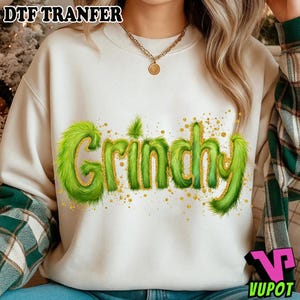 May include: Cream-coloured sweatshirt featuring the word "Grinchy" in green, furry lettering with gold glitter accents. The top has a crew neck and long sleeves. The text "DTF TRANFER" is at the top left.