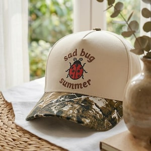 May include: A beige baseball cap with a camouflage brim. The cap features the embroidered words "sad bug summer" with a red and black ladybug design. The cap is sitting on a white cloth.