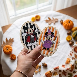 Könnte beinhalten: Zwei ovale, bestickte Aufnäher mit gewelltem Rand. Ein Aufnäher zeigt Mickey Maus als Vampir vor einem schwarz-weiß gestreiften Hintergrund. Der andere zeigt Minnie Maus als Hexe vor einem orange-weiß gestreiften Hintergrund.
