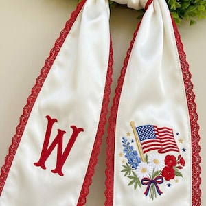 May include: White fabric sash with red lace trim. One sash features a large red "W" and the other has an embroidered American flag with flowers. The design is patriotic and suitable for festive occasions.