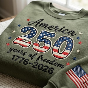 Op de afbeelding: Olijfgroene sweatshirt met geborduurde tekst en afbeeldingen. Het ontwerp bevat het woord "America" boven het getal "250" in rood, wit en blauw, en de tekst "years of freedom 1776-2026".