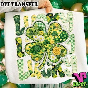 Op de afbeelding: Witte stof met een groot, gedetailleerd klaverbladontwerp. Het klaverblad is versierd met groene, gele en witte bloemaccenten en omgeven door een parelachtige rand. Het woord "LUCKY" is geschreven in groen en geel gekleurde letters.