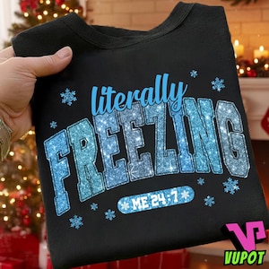 May include: Black sweatshirt with the text "literally FREEZING" in blue glitter lettering, surrounded by snowflakes. The phrase "ME 24:7" is below the main text. A cozy winter-themed apparel item.