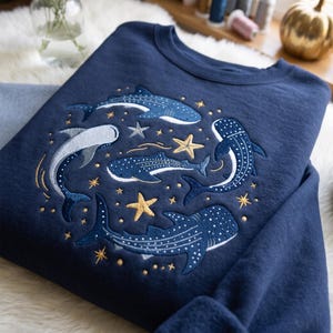 Pode incluir: Uma camisola azul-marinho de gola redonda com um design bordado de quatro tubarões-baleia a nadar num padrão circular. O bordado inclui tubarões-baleia brancos e azuis, estrelas-do-mar douradas e prateadas e pequenas estrelas douradas.