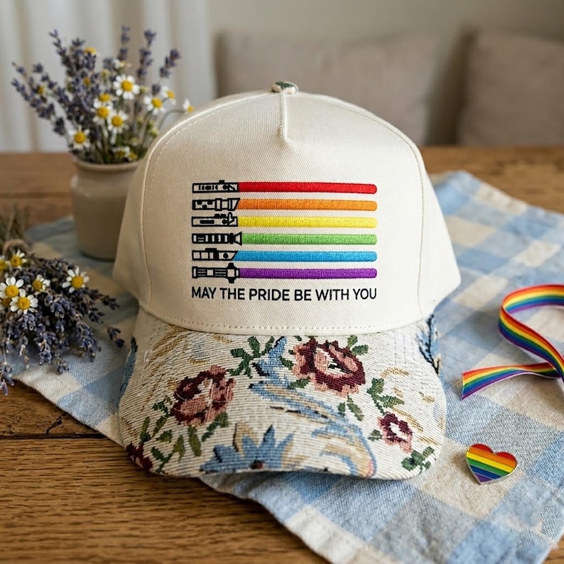May include: A white baseball cap with a floral patterned brim. The cap features a rainbow design with the text "MAY THE PRIDE BE WITH YOU". A rainbow ribbon and heart-shaped pin are also visible.