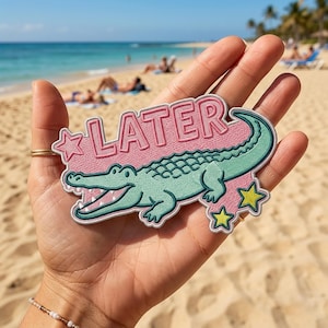 May include: An embroidered patch featuring a light green alligator positioned below the pink text LATER. The design includes a pink star on the left and two yellow stars on the bottom right, all outlined in white thread against a pink background.