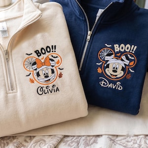 Könnte beinhalten: Ein cremefarbenes Sweatshirt mit Viertelreißverschluss, bestickt mit einer Mumien-Minnie-Maus und dem Namen Olivia, sowie ein marineblaues Sweatshirt mit einer Mumien-Mickey-Maus und dem Namen David, beide mit dem Text BOO!! und Fledermäusen.