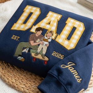 May include: Navy blue sweatshirt with embroidered "DAD" in yellow gingham and a graphic of a father and child on a skateboard. The sleeve is embroidered with the name "James". The sweatshirt also has the text "EST. 2026".