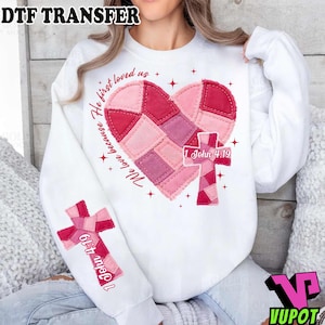 以下が含まれることがあります： ピンクと赤の色合いのパッチワークのハートと十字架のデザインが特徴の白いスウェットシャツ。「He first loved us, We love because」と「1 John 4:19」の文字がプリントされています。袖には十字架のデザインがあります。