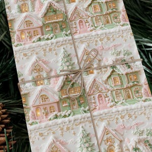 May include: A white gift wrap with a repeating pattern of pink and green houses with snow-covered roofs and lit windows. The houses are surrounded by a white border with gold accents.