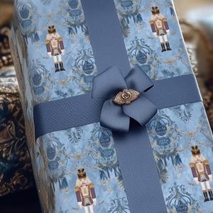 Op de afbeelding: Een ingepakte geschenkdoos met blauw inpakpapier met een patroon van notenkrakersoldaten en sierlijke ontwerpen. Een breed blauw lint en strik met een goudkleurige versiering beveiligen het pakket, wat een feestelijke presentatie creëert.
