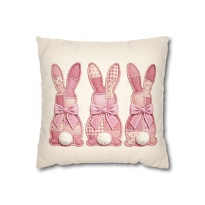 May include: A square, cream-colored decorative pillow featuring three pink bunny appliqués. Each bunny has patchwork ears, a bow, and a fluffy white tail. The bunnies are arranged vertically on the pillow.