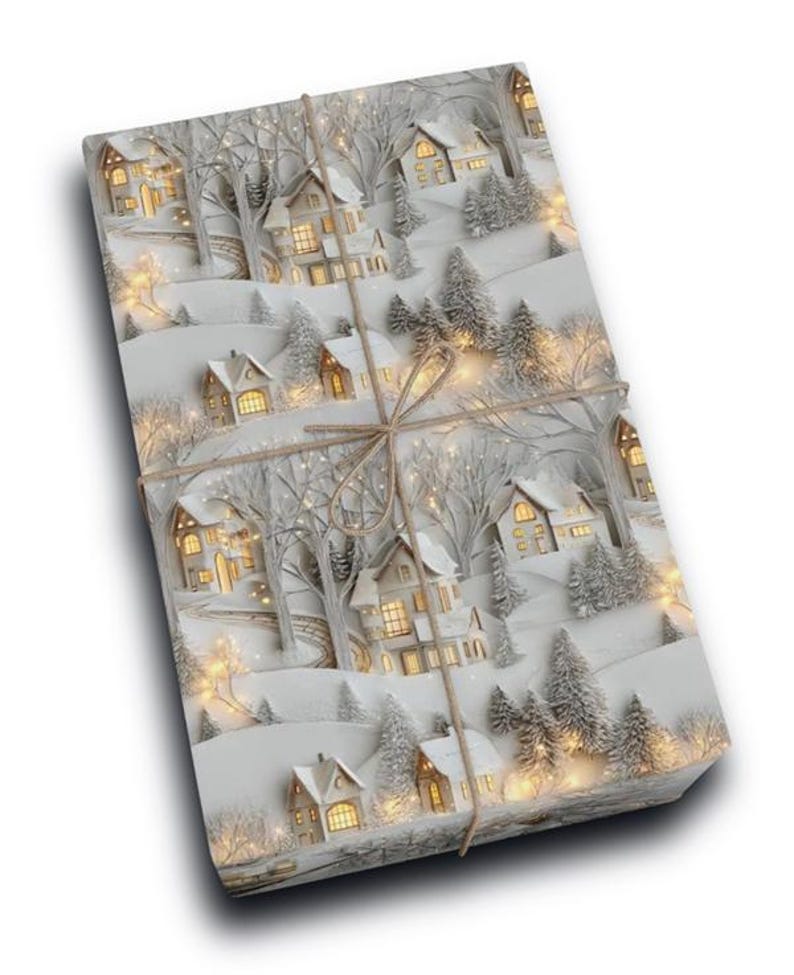 May include: A rectangular gift box wrapped in paper featuring a winter village scene with snow-covered houses, bare trees, and evergreen pines. The design includes warm yellow glowing lights in the windows and is tied with a thin brown twine string.