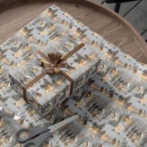 May include: A rectangular gift box wrapped in paper featuring a repeating pattern of snow-covered houses and trees with glowing yellow windows. The box is tied with a brown satin ribbon bow. A roll of clear tape and grey scissors are placed nearby.