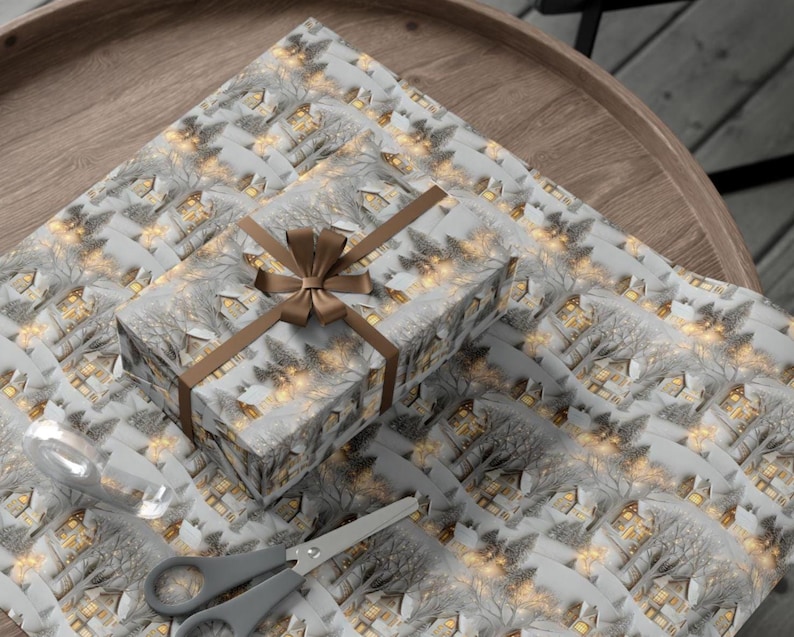 May include: A rectangular gift box wrapped in paper featuring a repeating pattern of snow-covered houses and trees with glowing yellow windows. The box is tied with a brown satin ribbon bow. A roll of clear tape and grey scissors are placed nearby.