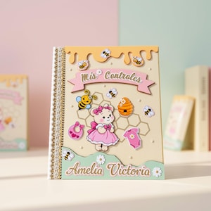May include: A rectangular scrapbook with a cream cover featuring a honey drip design at the top, a pink banner with the text "Mis Controles," a cartoon bear in a pink dress, a beehive, bees, a bib, and a onesie. The name "Amelia Victoria" is printed at the bottom.