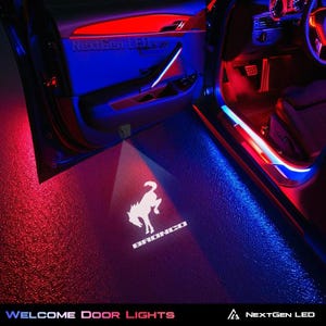 May include: A pair of red and blue LED welcome door lights project the Ford Bronco logo onto the ground. The lights are installed in a car door and illuminate the area around the door when the door is opened. The lights are labelled "NextGen LED" and "Welcome Door Lights".