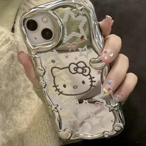May include: A silver phone case with a Hello Kitty design. The case is decorated with rhinestones and stars. The case is clear and shows the phone's camera lens.