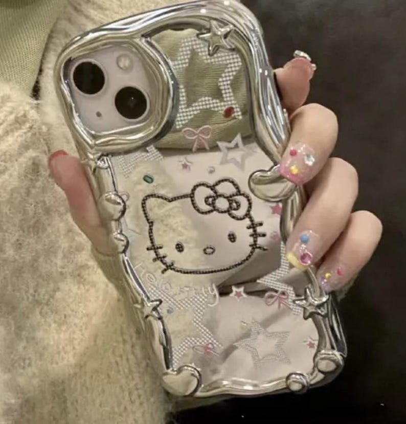 May include: A silver phone case with a Hello Kitty design. The case is decorated with rhinestones and stars. The case is clear and shows the phone's camera lens.