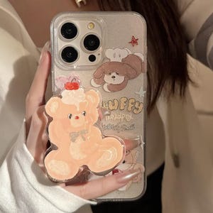 Peut inclure: Une coque de téléphone transparente avec un fond pailleté et des autocollants d'animaux de dessin animé. La coque présente un chien brun portant un chapeau de chef, un ours rose avec une cerise sur le dessus et le texte "LUFFY" en police rose et blanche.