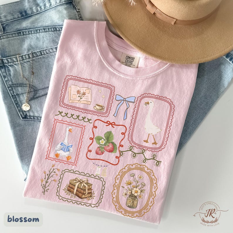 May include: A light pink t-shirt featuring framed illustrations of a goose, strawberries, books, and flowers. The shirt is adorned with a blue bow and the word "blossom". A tan hat and blue jeans are in the background.