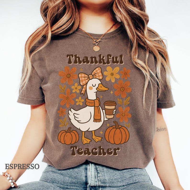 May include: Espresso-colored t-shirt featuring a cartoon goose with a scarf and coffee cup, surrounded by flowers and pumpkins. The text reads "Thankful Teacher". A fall-themed design.