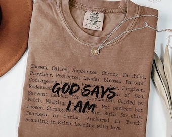May include: Espresso-colored t-shirt with the text "GOD SAYS I AM" and a list of affirmations. A silver necklace with a pendant rests on the shirt. The shirt has a Comfort Colors tag.