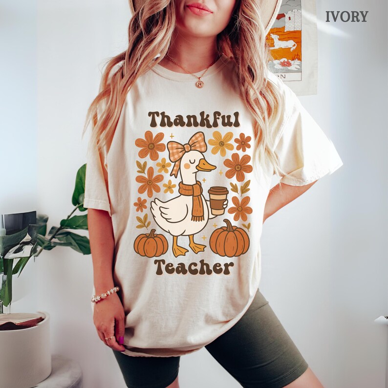 May include: Ivory-colored t-shirt with a fall-themed graphic. The design features a cartoon goose wearing a scarf and holding a coffee cup, surrounded by flowers and pumpkins. Text reads "Thankful Teacher".