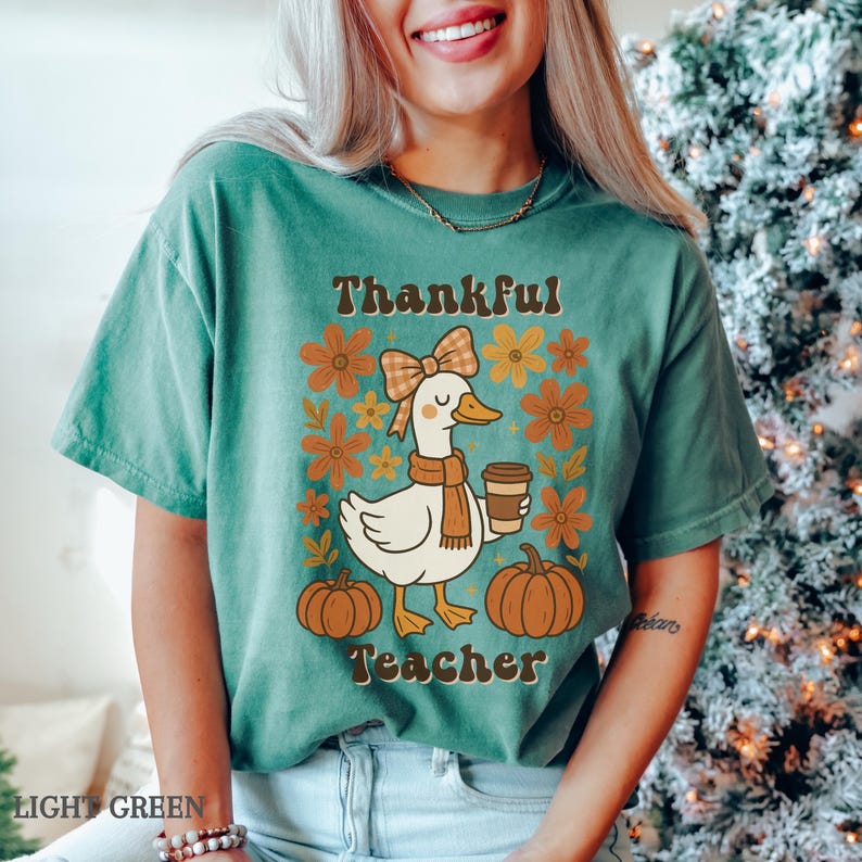 May include: Light green t-shirt with a graphic featuring a cartoon duck wearing a scarf and holding a coffee cup. The text "Thankful Teacher" is above the duck, with pumpkins and flowers surrounding the duck.