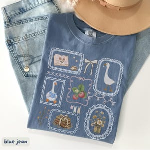 May include: A blue t-shirt with a whimsical design featuring framed illustrations of a goose, strawberries, books, and flowers. The shirt is paired with light blue jeans and a tan hat, creating a casual, aesthetic look. A gold necklace is also visible.