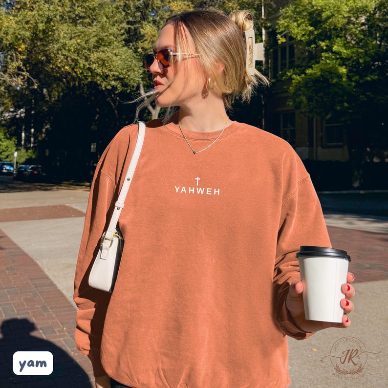 May include: Rust-colored sweatshirt with the word "YAHWEH" and a cross printed on the front. The sweatshirt is paired with a white purse and sunglasses. A person is holding a white coffee cup with a black lid.