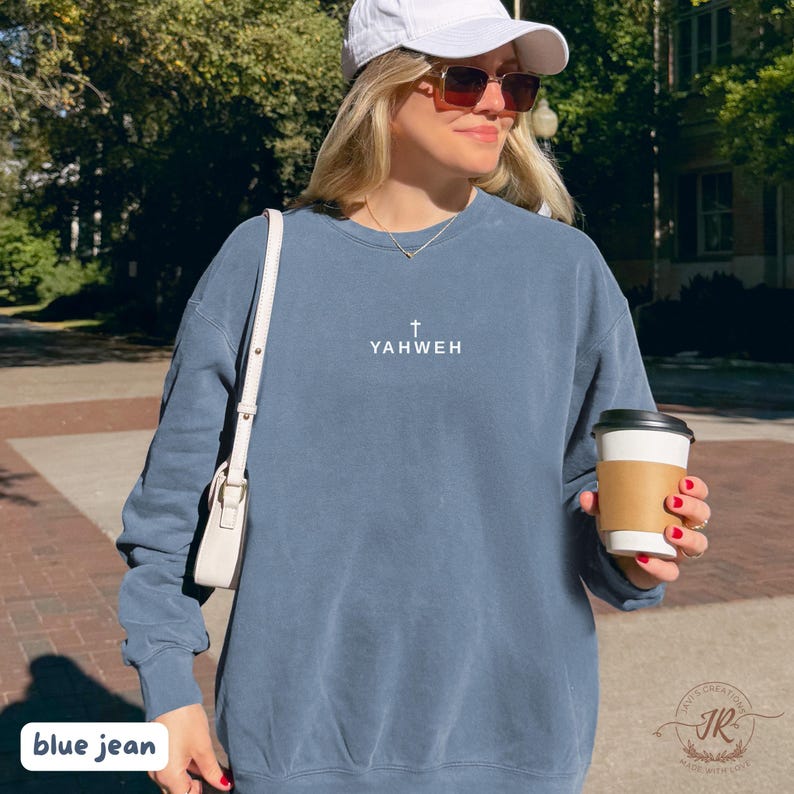 May include: Blue jean colored sweatshirt with the word "YAHWEH" and a cross symbol printed on the front. The sweatshirt is a solid color and has long sleeves. The person in the image is wearing a white cap and sunglasses.