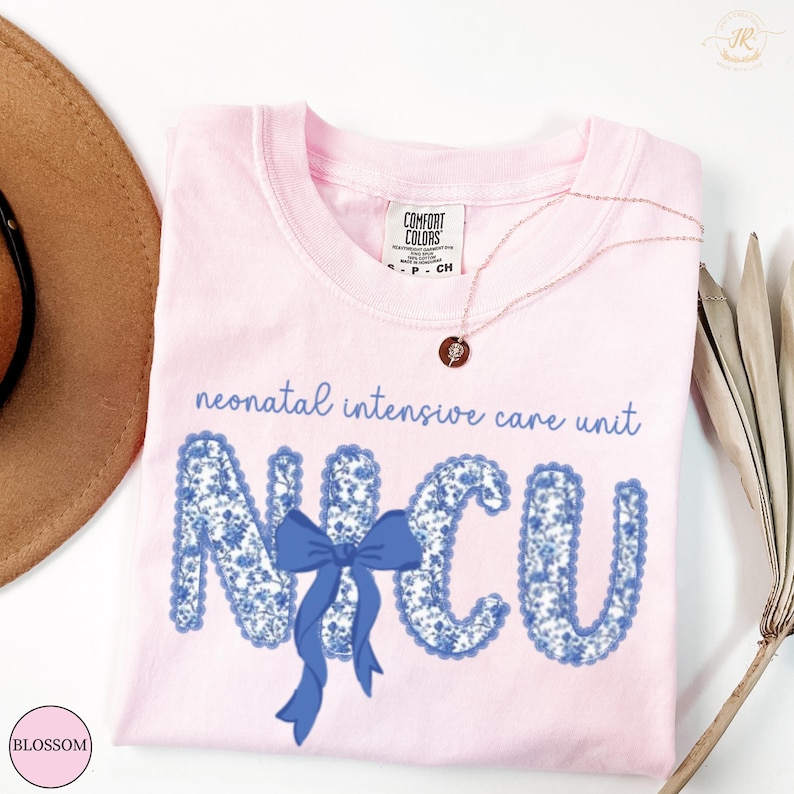 May include: A light pink t-shirt with the words "neonatal intensive care unit" and the letters "NICU" in a blue floral pattern. A blue bow is centered between the letters. A gold necklace with a pendant is on the shirt.