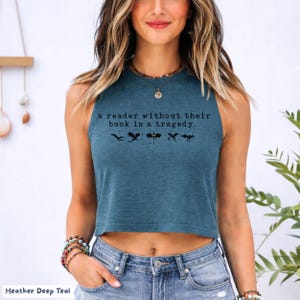 May include: A teal crop top with the text "a reader without their book is a tragedy." The shirt features black silhouette graphics of dragons. The bottom of the shirt is visible, along with the model's blue jeans and bracelets.