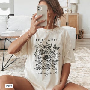 May include: Ivory-coloured t-shirt with a floral and butterfly design, and the text "IT IS WELL with my soul" in black. The shirt has a relaxed fit and short sleeves. The style is casual and comfortable.