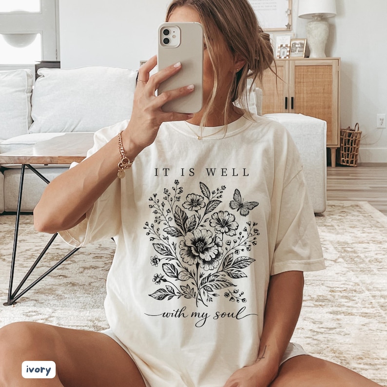 May include: Ivory-coloured t-shirt with a floral and butterfly design, and the text "IT IS WELL with my soul" in black. The shirt has a relaxed fit and short sleeves. The style is casual and comfortable.