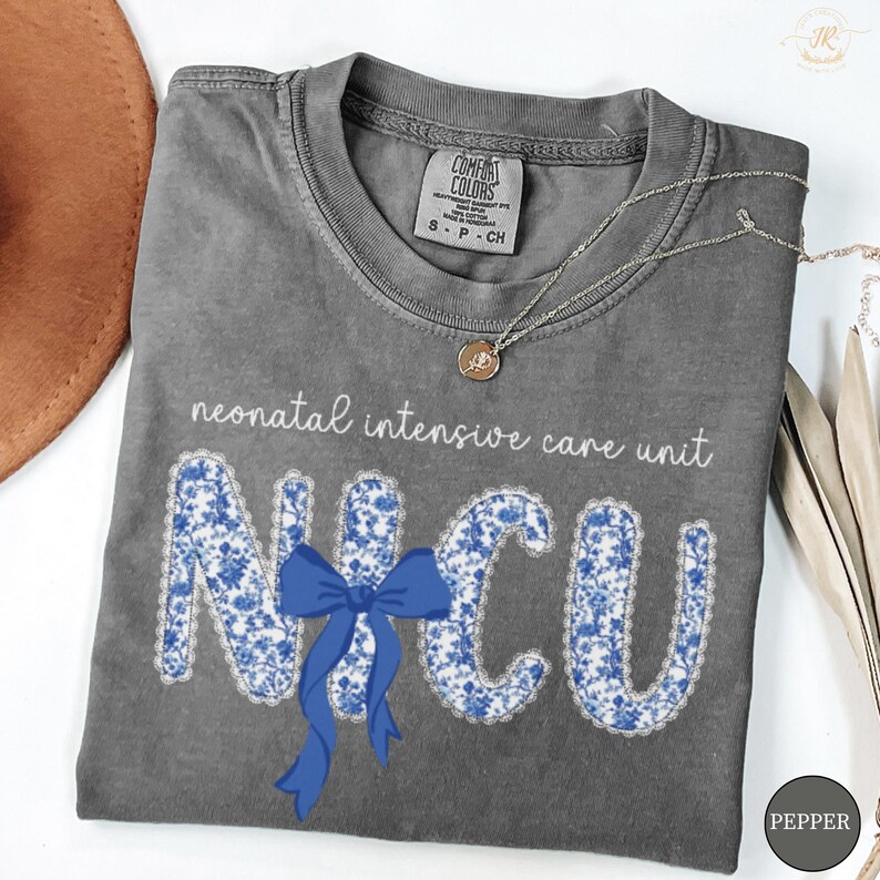 May include: A gray t-shirt with the words "neonatal intensive care unit" and the letters "NICU" in a blue floral pattern. A blue bow is centered below the letters. A silver necklace with a small pendant is also visible.