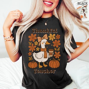 May include: Black t-shirt with a fall-themed graphic. The design features a cartoon goose wearing a scarf and holding a coffee cup, surrounded by flowers and pumpkins. The text reads "Thankful Teacher".