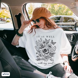 May include: White crew neck t-shirt with a black floral and butterfly design, and the words "IT IS WELL with my soul". The shirt is a casual style.