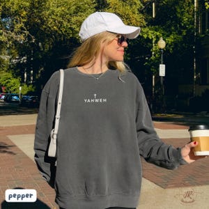 May include: A dark gray sweatshirt with the word "YAHWEH" printed in white, along with a small cross. The sweatshirt is worn by a person wearing a white baseball cap and sunglasses. The word "pepper" is in a white box in the lower left corner.