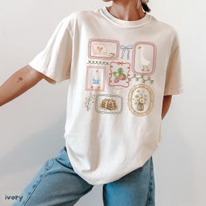 May include: Ivory-colored t-shirt featuring a collection of framed illustrations. The illustrations include a goose, strawberries, flowers, books, and bows. The shirt has a relaxed fit and is made of a soft material. The word "ivory" is printed at the bottom left.