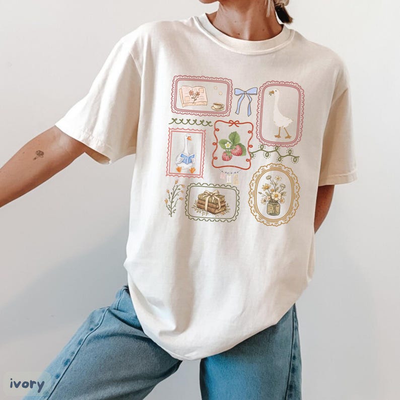May include: Ivory-colored t-shirt featuring a collection of framed illustrations. The illustrations include a goose, strawberries, flowers, books, and bows. The shirt has a relaxed fit and is made of a soft material. The word "ivory" is printed at the bottom left.