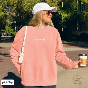 May include: Peach-colored sweatshirt with the word "YAHWEH" and a cross printed on the front. The sweatshirt is worn with a white cap and sunglasses. A white purse is slung over the shoulder. The image is taken outdoors.