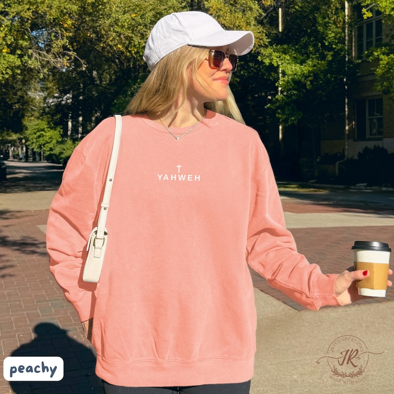 May include: Peach-colored sweatshirt with the word "YAHWEH" and a cross printed on the front. The sweatshirt is worn with a white cap and sunglasses. A white purse is slung over the shoulder. The image is taken outdoors.